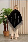 Woman wearing a black poncho with fringe details, standing indoors next to a plant.