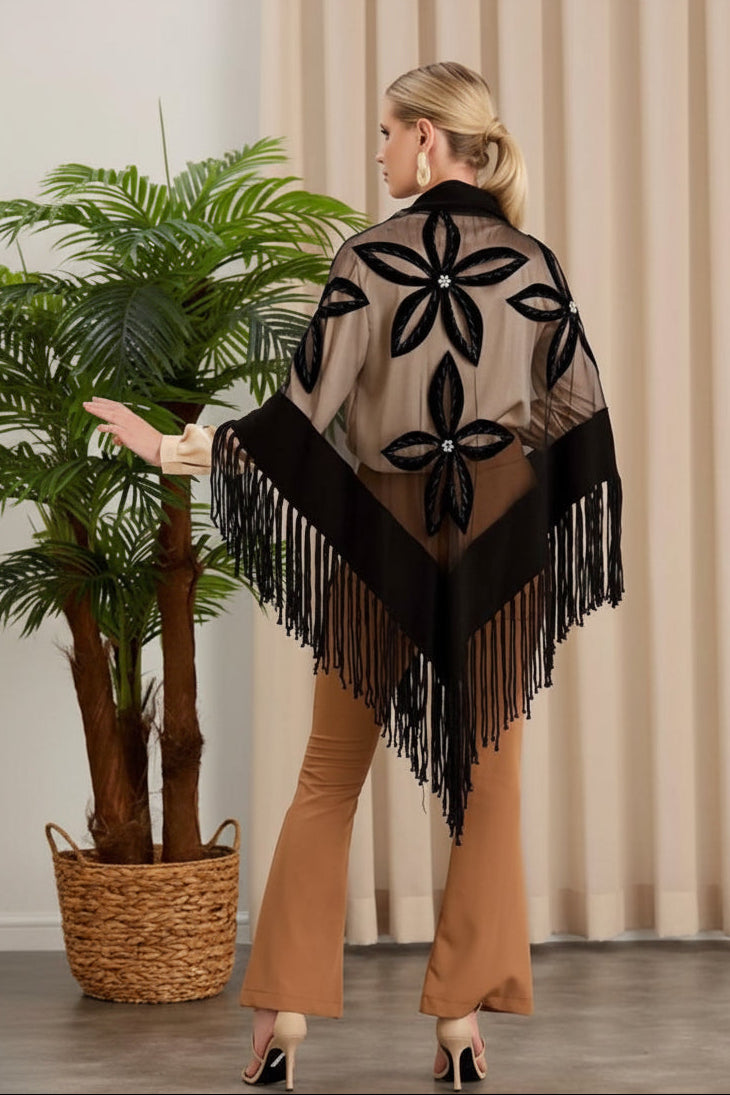 Woman wearing a patterned shawl with fringe, standing indoors next to a plant.