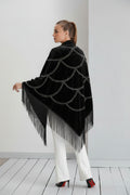Woman wearing a black poncho with fringe details on a white floor and light gray wall.