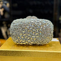 A heavily embellished rectangular evening clutch bag covered in silver rhinestones and iridescent marquise crystals, displayed on a textured gold box against a dark, blurred background.