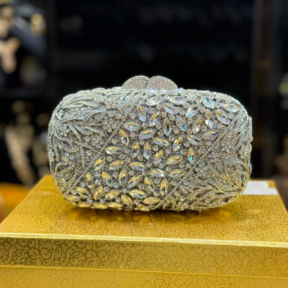 A heavily embellished rectangular evening clutch bag covered in silver rhinestones and iridescent marquise crystals, displayed on a textured gold box against a dark, blurred background.