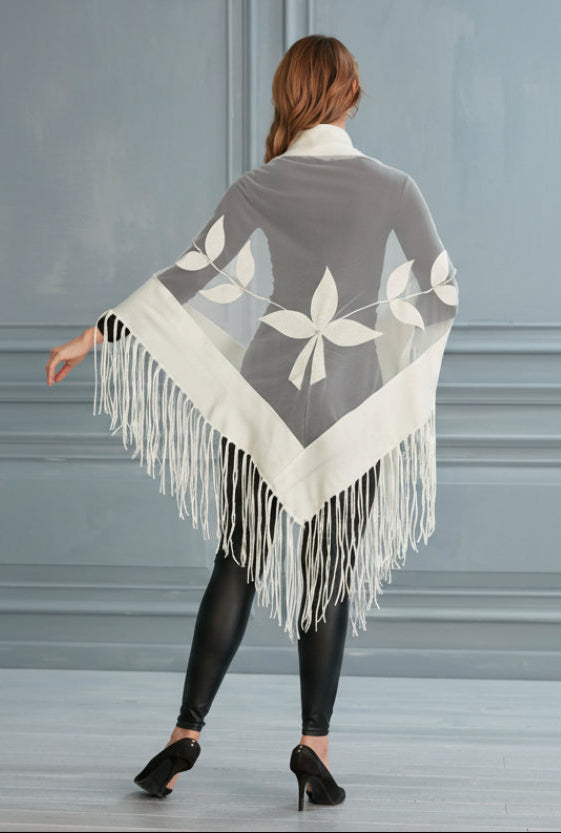 Woman wearing a gray and white patterned poncho with fringes, standing against a plain background.